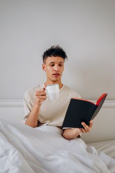 Young man reading a book while holding a coffee cup in a cozy bedroom setting.