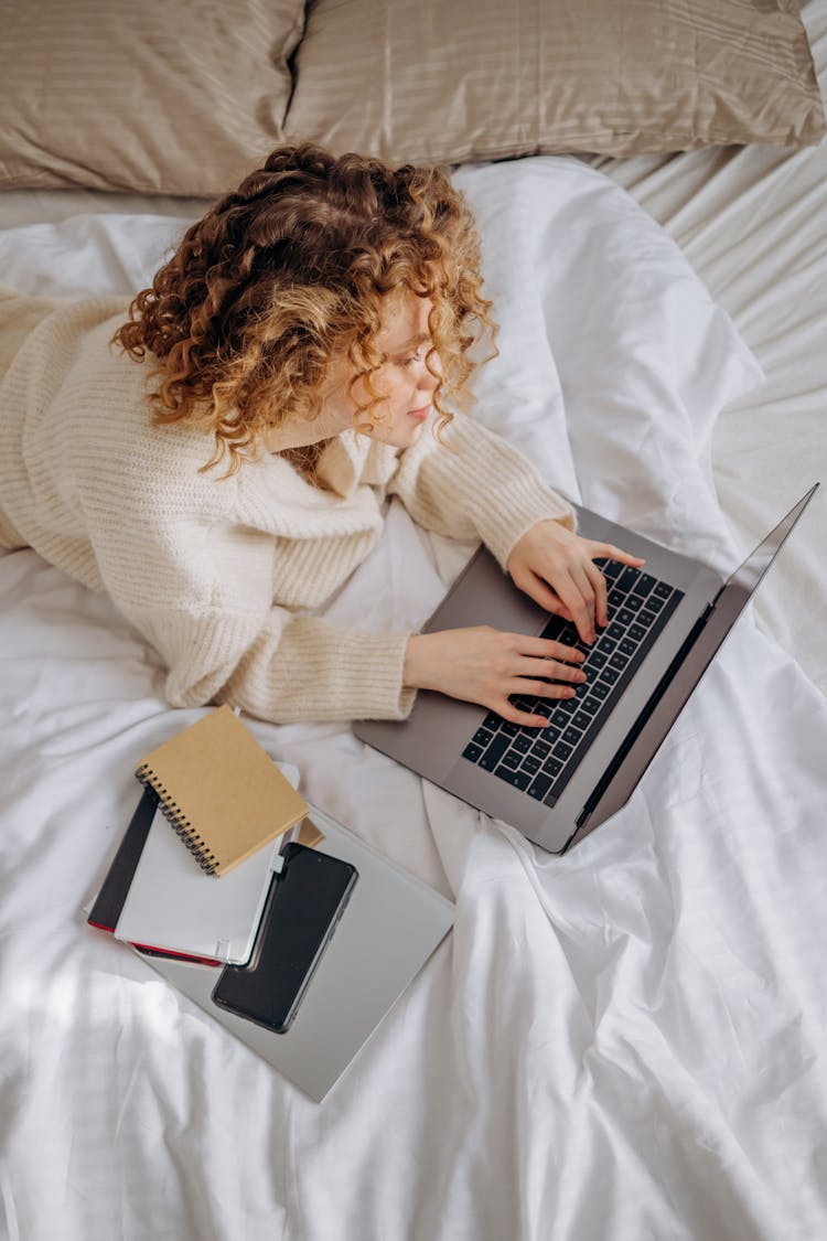 Woman In Beige Sweater Using Laptop On Bed In Prone Position