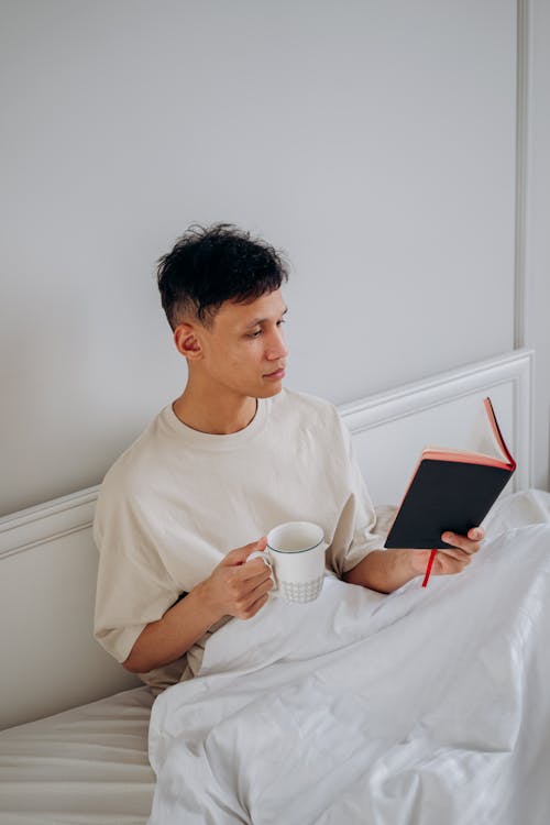 Free Man Sitting on the Bed while Holding a Cup of Coffee and Reading a Book Stock Photo