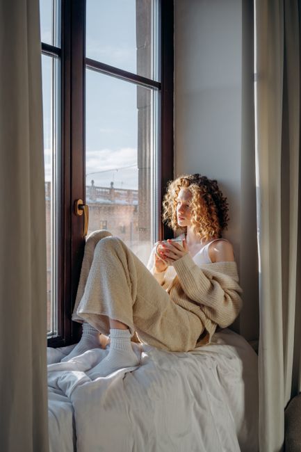 Woman enjoying a quiet morning with a drink by the window, embracing cozy knitwear.