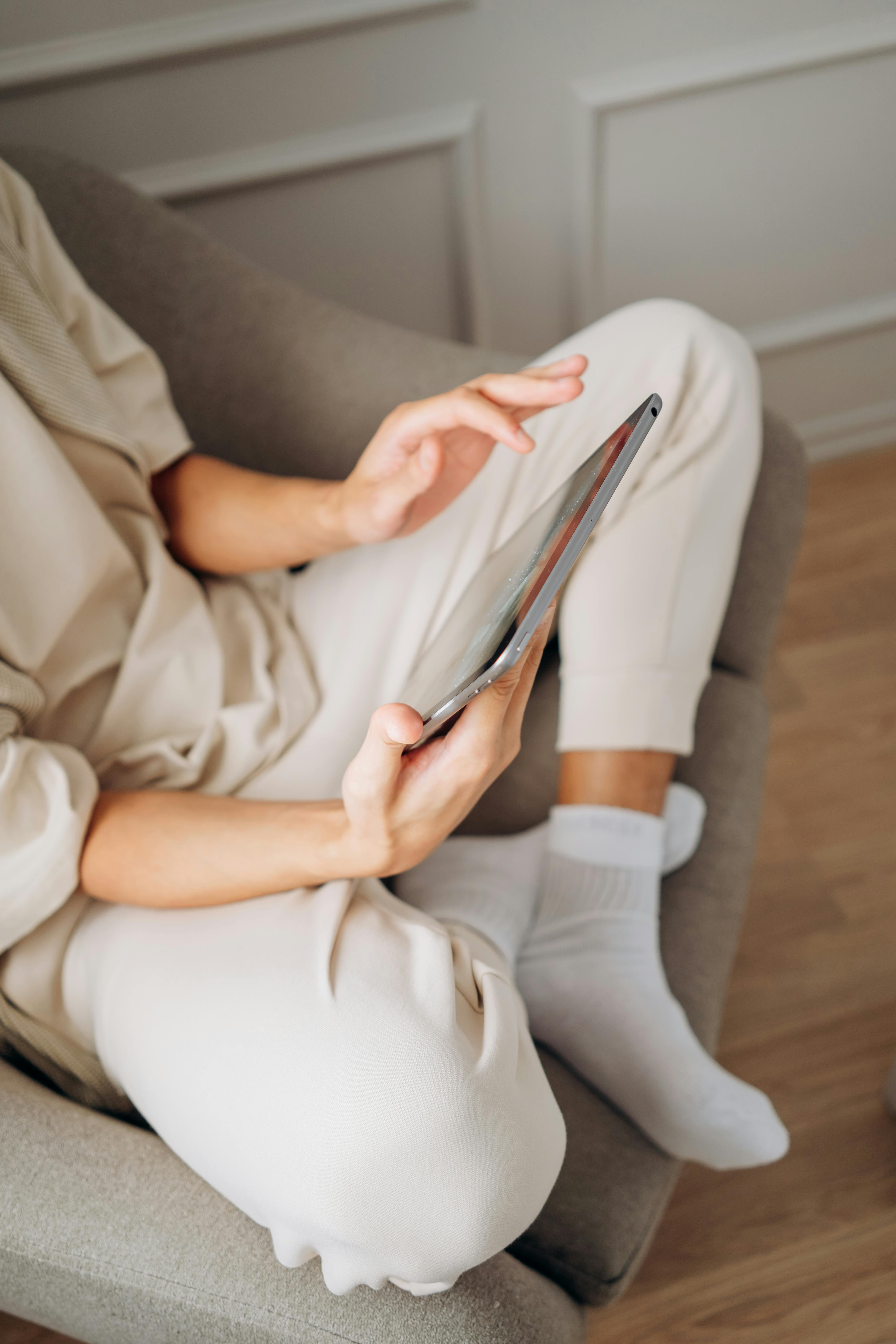 Free A person sits comfortably on a sofa using a tablet, depicting a relaxed lifestyle indoors. Stock Photo