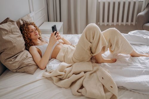 Free Cozy morning scene of a woman relaxing on bed with a smartphone. Stock Photo