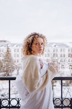 Caucasian woman with curly hair in a beige sweater holding coffee on a winter balcony.