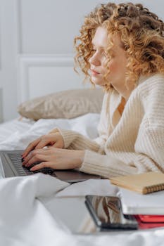 A young woman with curly hair works on a laptop, laying comfortably in a cozy bedroom setting.
