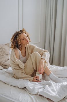 A woman enjoys a relaxing morning with a hot drink in a minimalist bedroom setting.