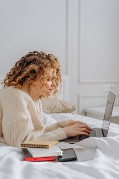 Woman with curly hair typing on a laptop while relaxing in a cozy, knit sweater on a bed.