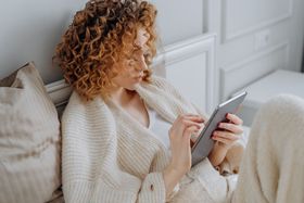 A woman relaxing on a bed, using a digital tablet while wearing a cozy knitted sweater.