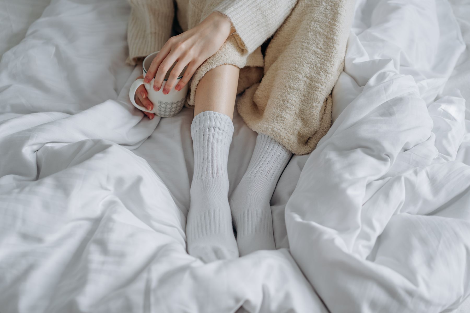 Feet in white wool socks on a bed under a white duvet, a mug in hand