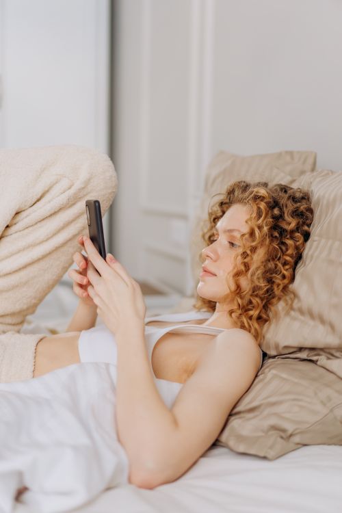 Free Young woman with curly hair lying in bed, using smartphone, enjoying leisure time indoors. Stock Photo