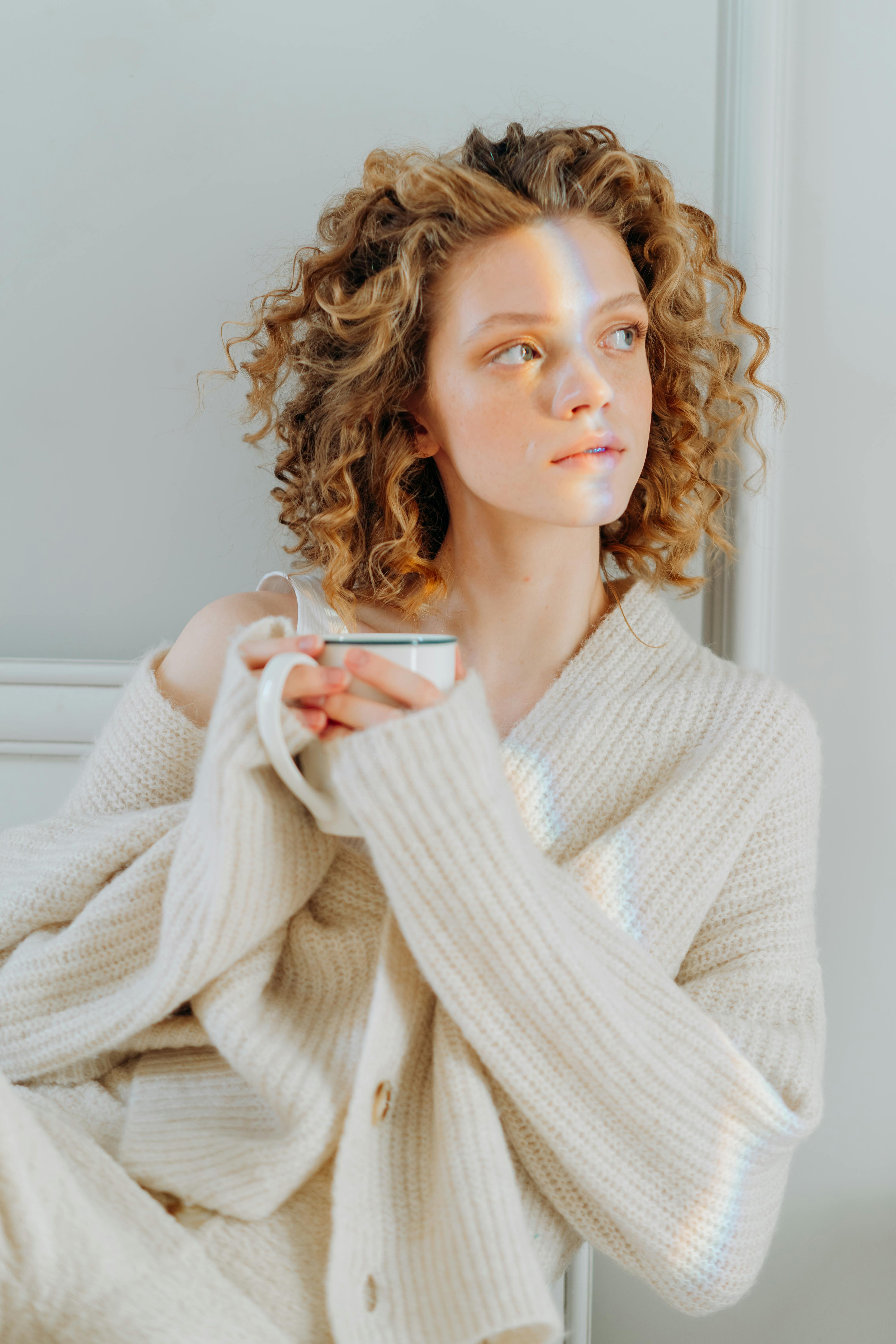 Woman with curly hair in a cream sweater holding a mug, bathed in gentle light.