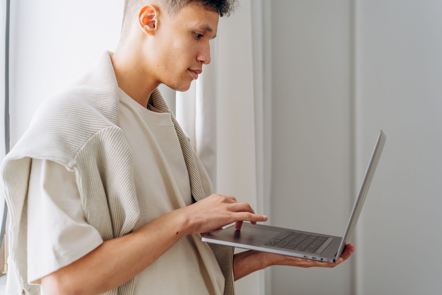 Stylish young man using a laptop indoors, focused on the screen during the day.