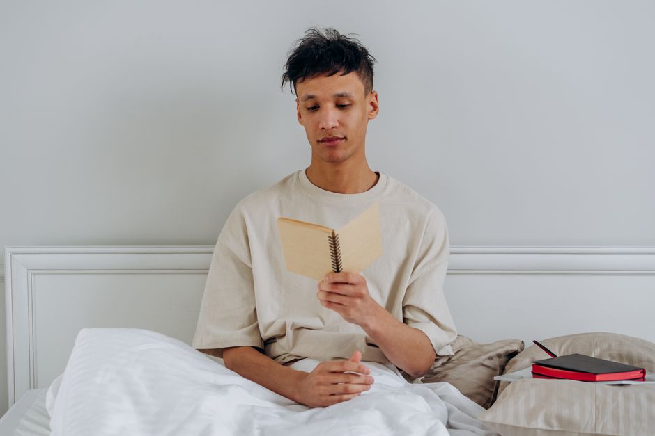 A young man sitting in bed reading with a notebook beside him, conveying relaxation and study. A young man sitting in bed reading with a notebook beside him, conveying relaxation and study.