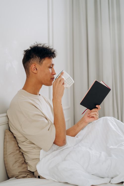 Free A young man sits in bed drinking from a mug while reading a book. Stock Photo