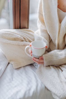 Relaxed scene of a person in a sweater holding a warm drink by the window.