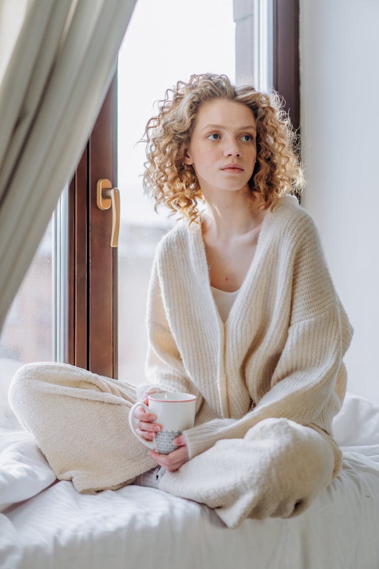 Woman In White Knit Sweater Holding White Ceramic Mug Sitting By A Glass Window