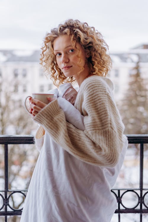 Woman in White Sweater Holding White Ceramic Mug Standing Near Metal Railings