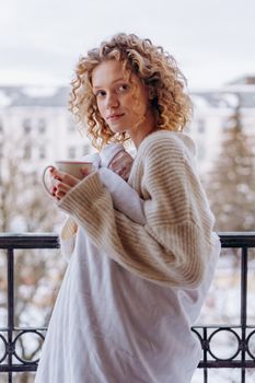 Young woman with curly hair stands on balcony holding coffee, wrapped in a cozy sweater.