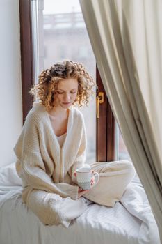 Woman enjoying a peaceful morning with a cup of coffee by a sunny window, wearing a cozy sweater.