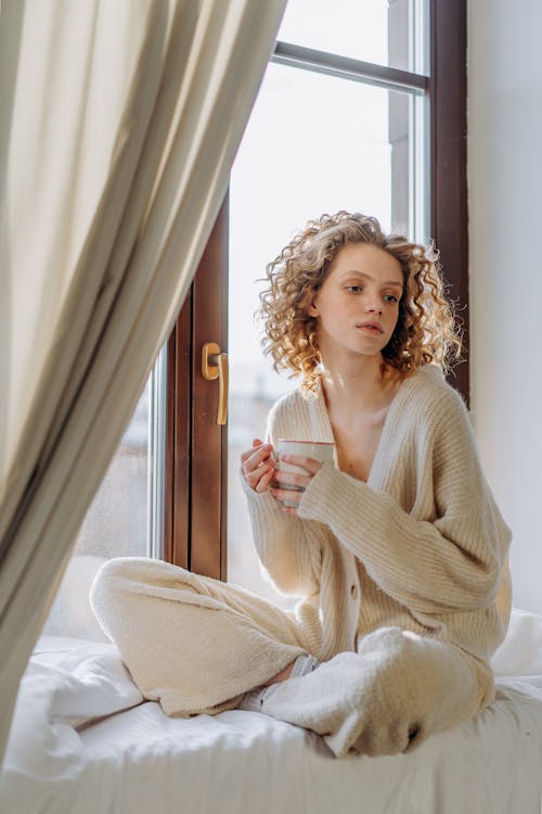 Woman in White Knit Sweater Sitting on White Linen by the Window