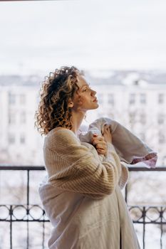 A woman stands on a balcony wrapped in a blanket, enjoying a peaceful winter morning.