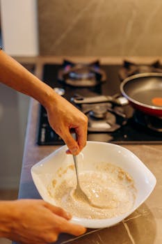 Close-up of hands mixing batter in a white bowl near a lit stove in a modern kitchen setting.
