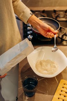 A person measures flour with a spoon next to a gas stove, ready to cook.