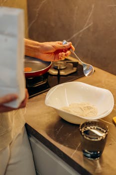 Person holding spoon with flour, preparing ingredients on a kitchen counter.
