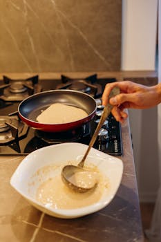 Close-up of pancake batter being ladled into a pan on a gas stove.