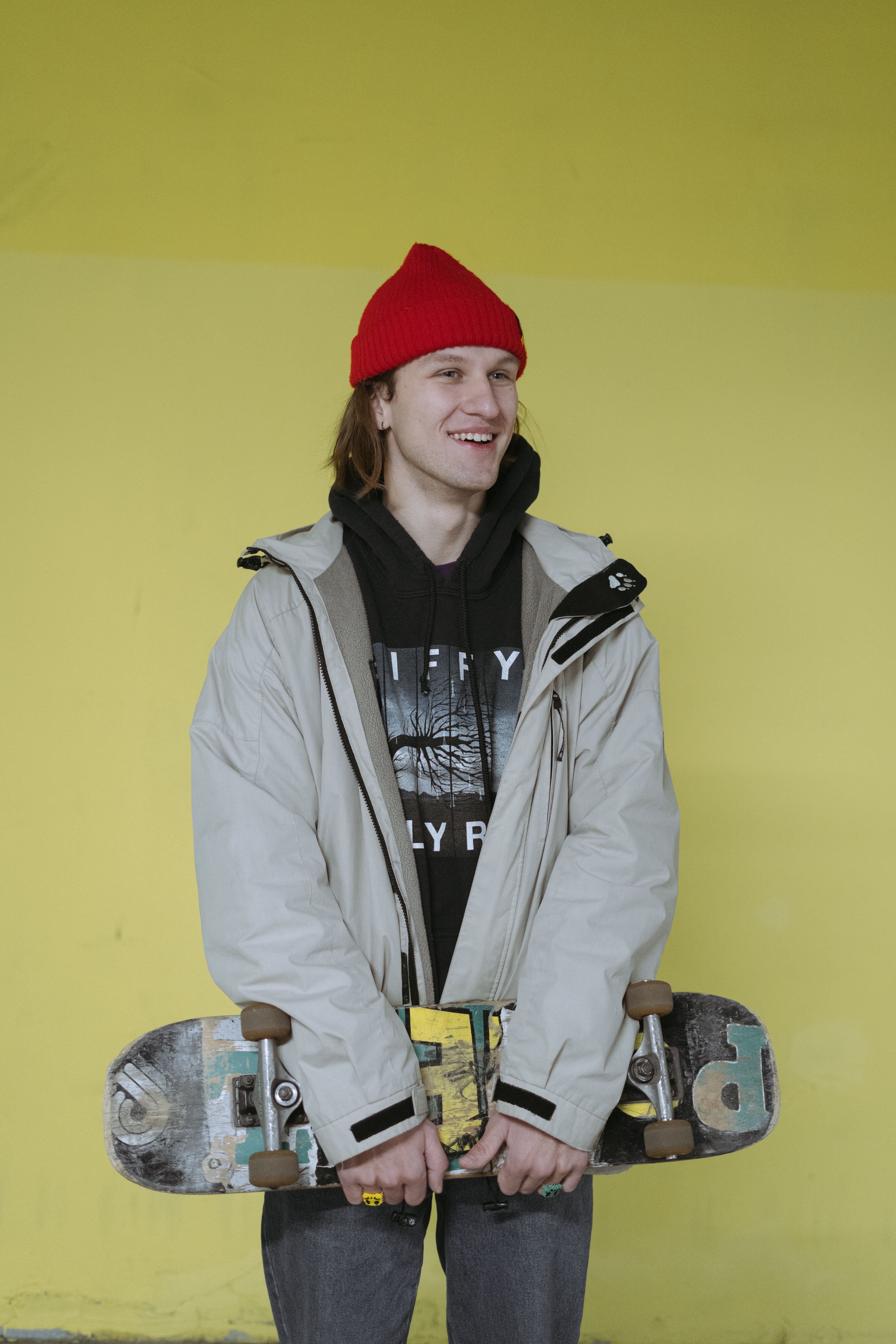 Young adult skater smiling in a red beanie holding a skateboard against a yellow wall.