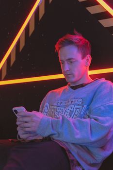 A young man in a gray sweater texting on a smartphone with vibrant neon lights.