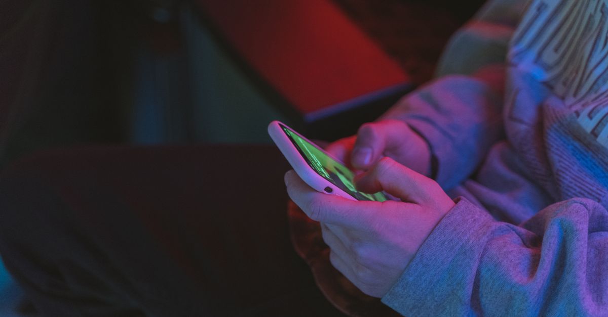 A person holding a smartphone with a Bible app open, sitting alone in a dimly lit room, illuminated only by the screen