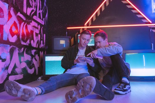 Two young men sitting in a neon-lit room interacting with a mobile phone surrounded by retro televisions.