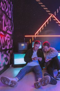 Two young men sitting in a neon-lit room with graffiti, enjoying technology with retro TVs.