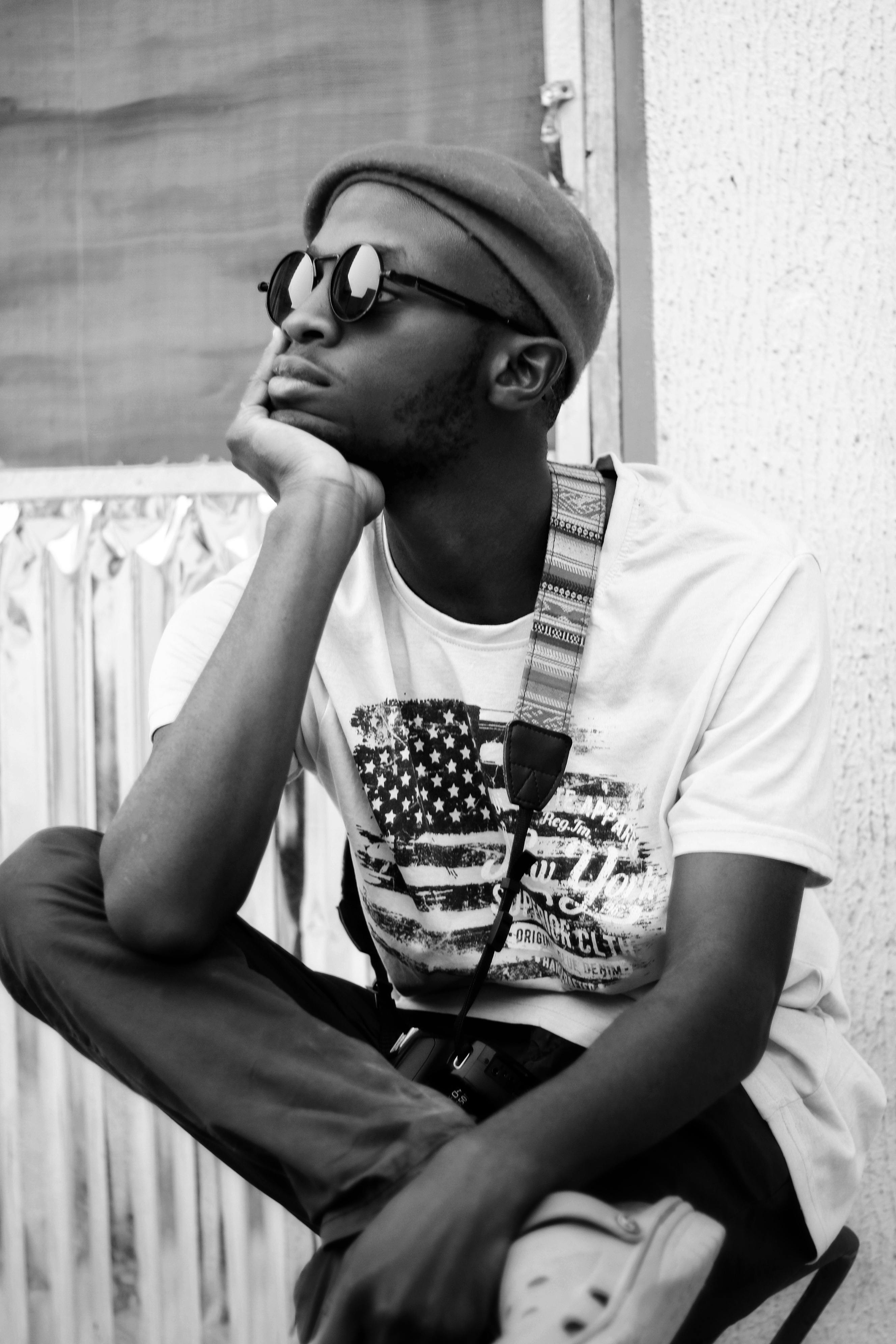 Black and white of trendy young African American male millennial in casual outfit and sunglasses sitting on chair with hand at chin in backyard and looking away pensively