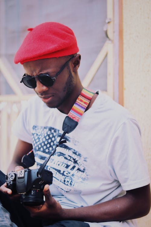 Concentrated young black male traveler in trendy hat and sunglasses setting up photo camera while standing near aged house on street