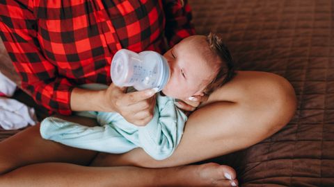 Crop anonymous mother feeding adorable newborn baby with bottle while sitting on bed with crossed legs in light bedroom at home