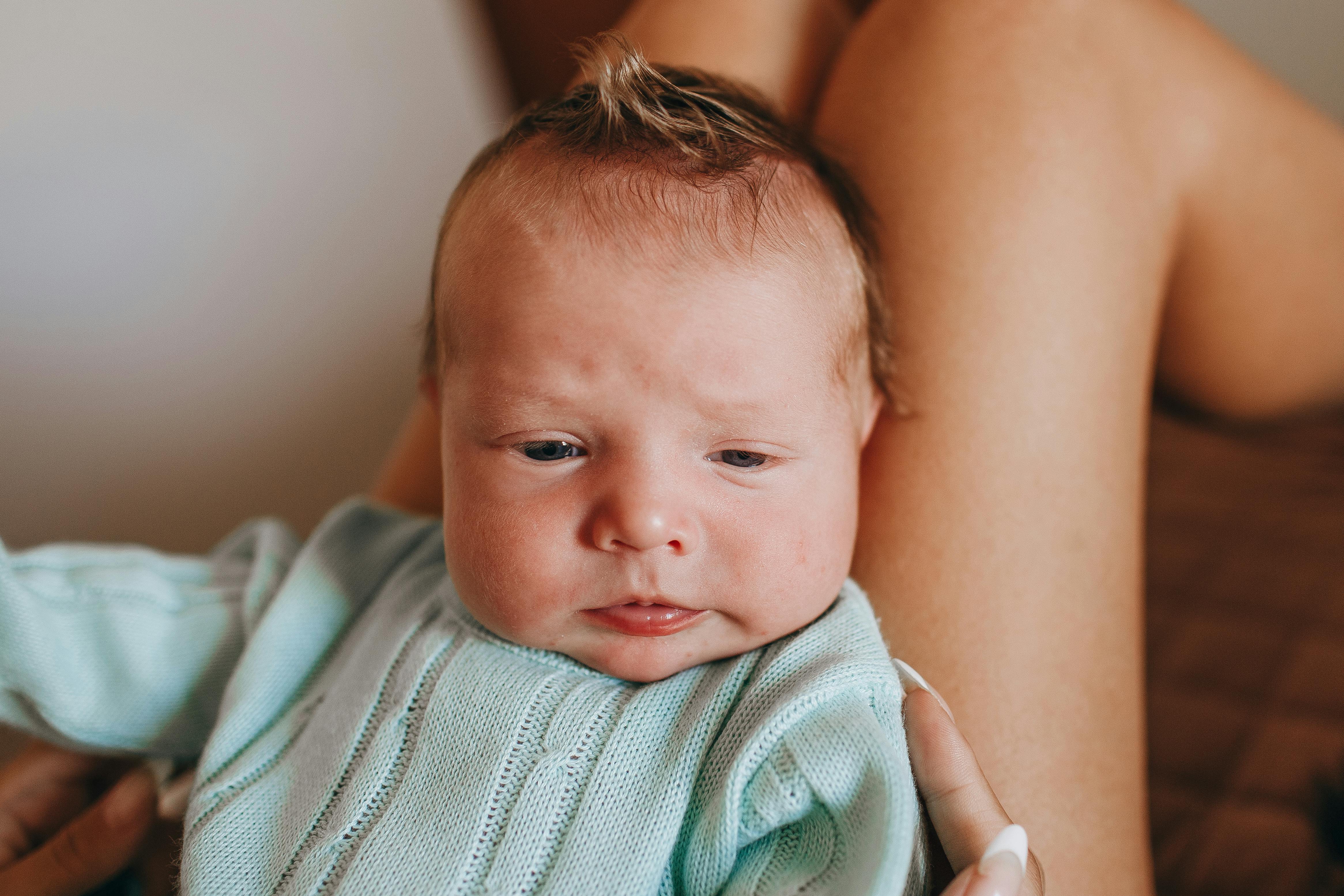 Crop unrecognizable mother with adorable little baby in wear on legs resting on comfortable bed in light bedroom at home