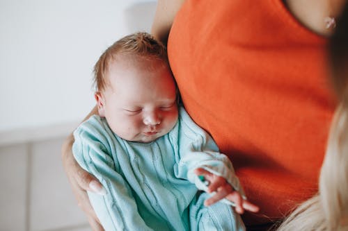 Crop anonymous mother with cute sleeping baby in warm wear in arms sitting in light room near wall at home