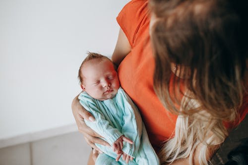 Crop anonymous mother with long hair with sleeping baby in arms sitting in light room near white wall at home