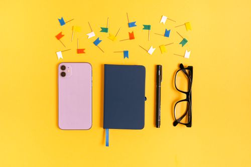 Free A vibrant flat lay of a smartphone, notebook, pen, eyeglasses, and colorful pins on a yellow background. Stock Photo