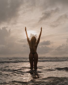 Back view of woman in bikini with arms raised standing in ocean at sunset.