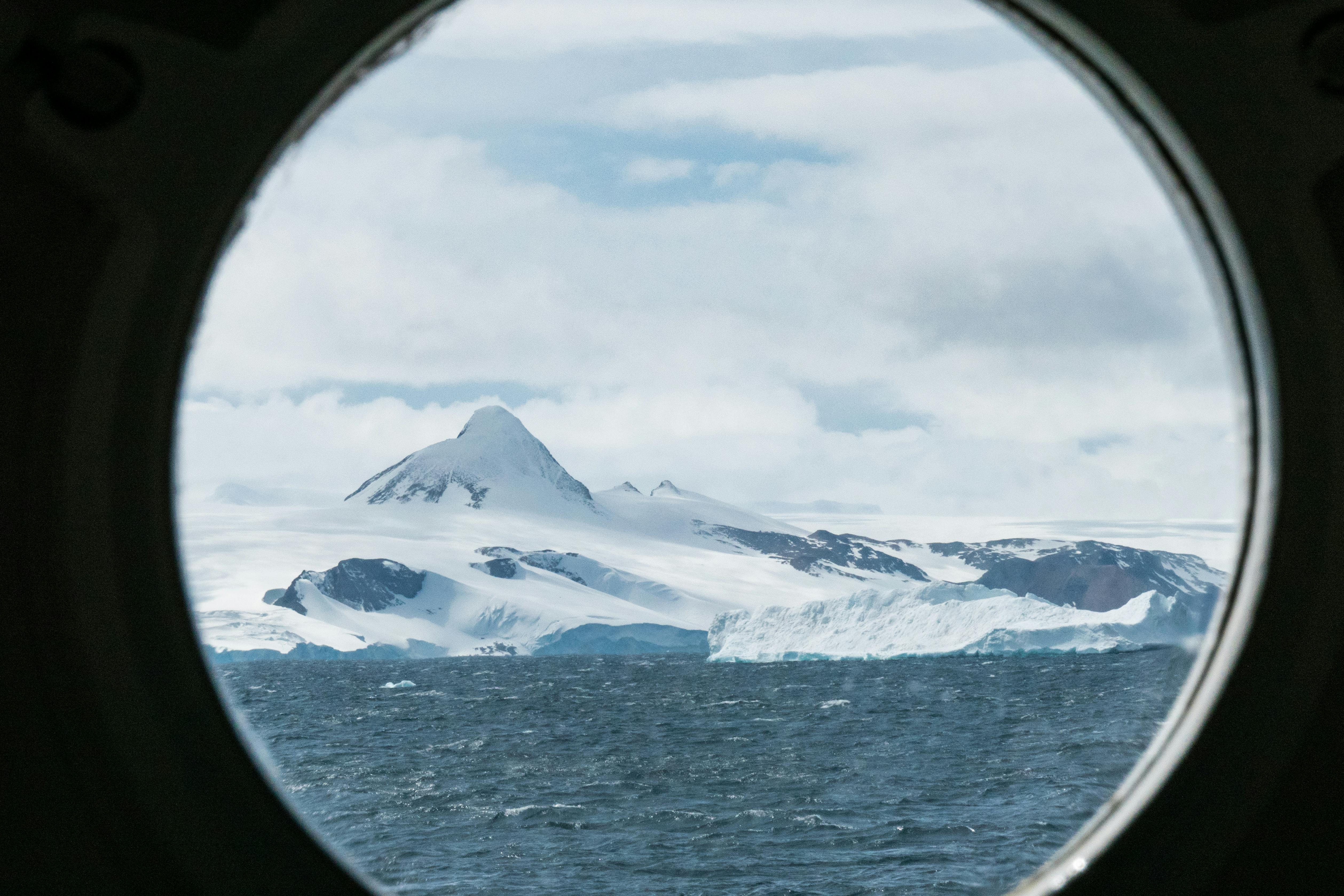 A stunning view of Antarctica's icy landscape through a ship's porthole, featuring snow-capped mountains and the open sea.