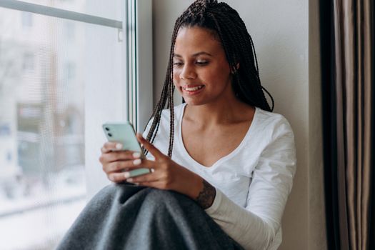 A woman sits by a window, smiling as she uses her smartphone, conveying a sense of relaxation and connectivity.