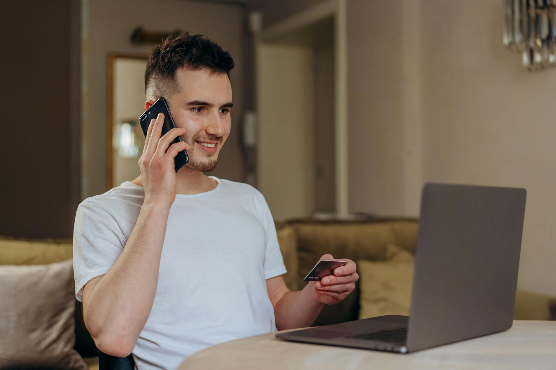 A young man smiles while shopping online using a phone and laptop, holding a credit card.