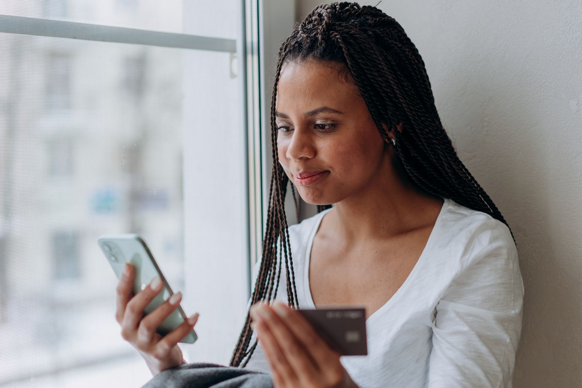 Young woman with braided hair using a smartphone and credit card by a window, smiling.