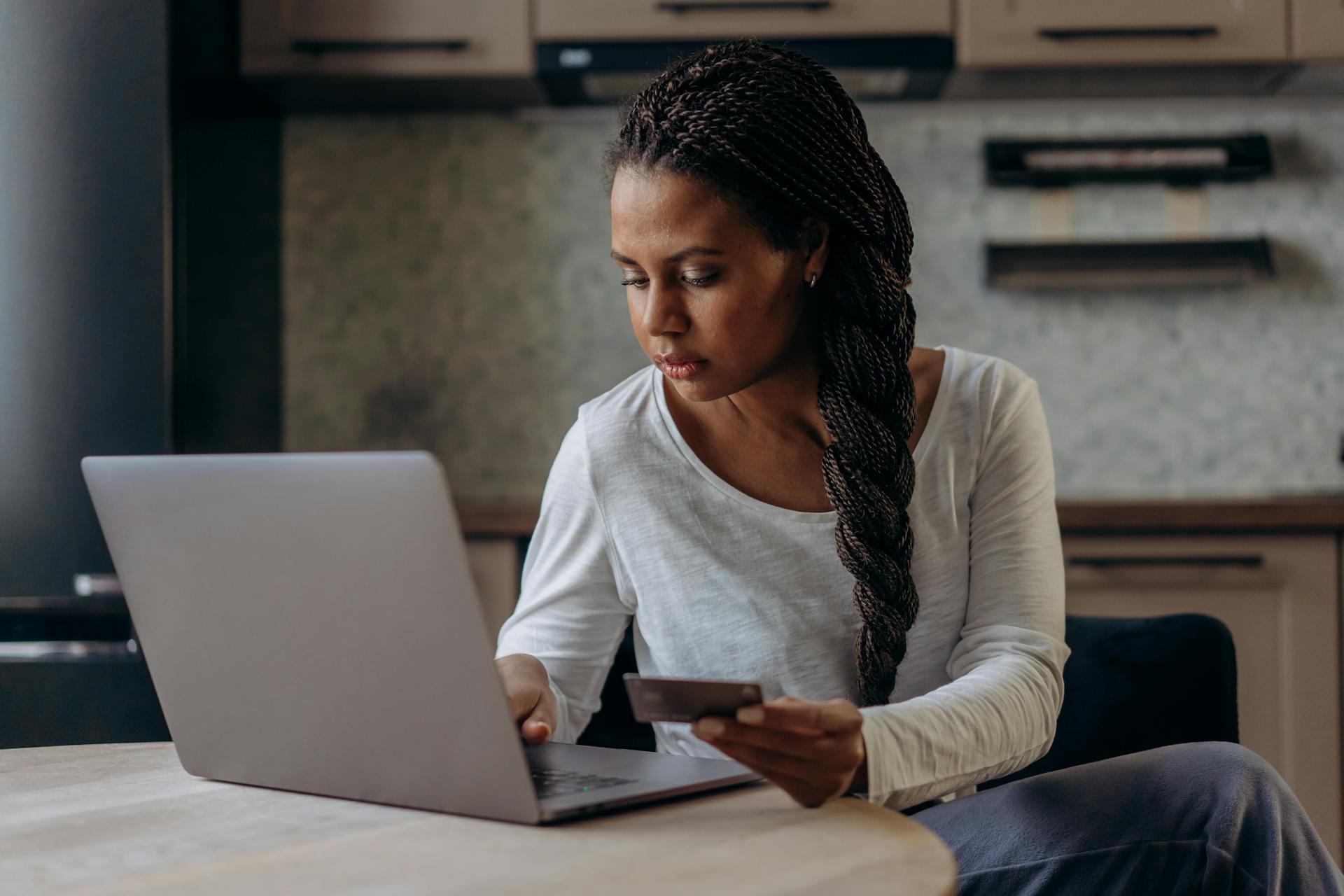 A woman with braided hair uses her laptop and credit card for online shopping indoors.