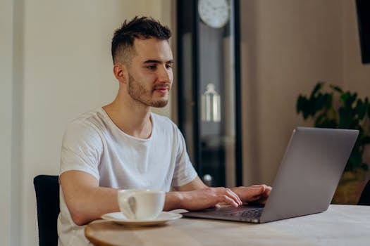 Adult man in a white shirt working on a laptop with a cup of coffee at a table indoors.