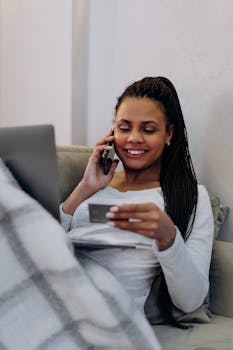 Smiling woman with smartphone and credit card shopping online while cozy at home.