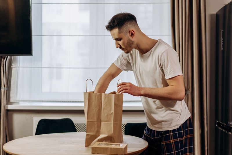 A young man is unpacking groceries from a paper bag onto a table inside his home.