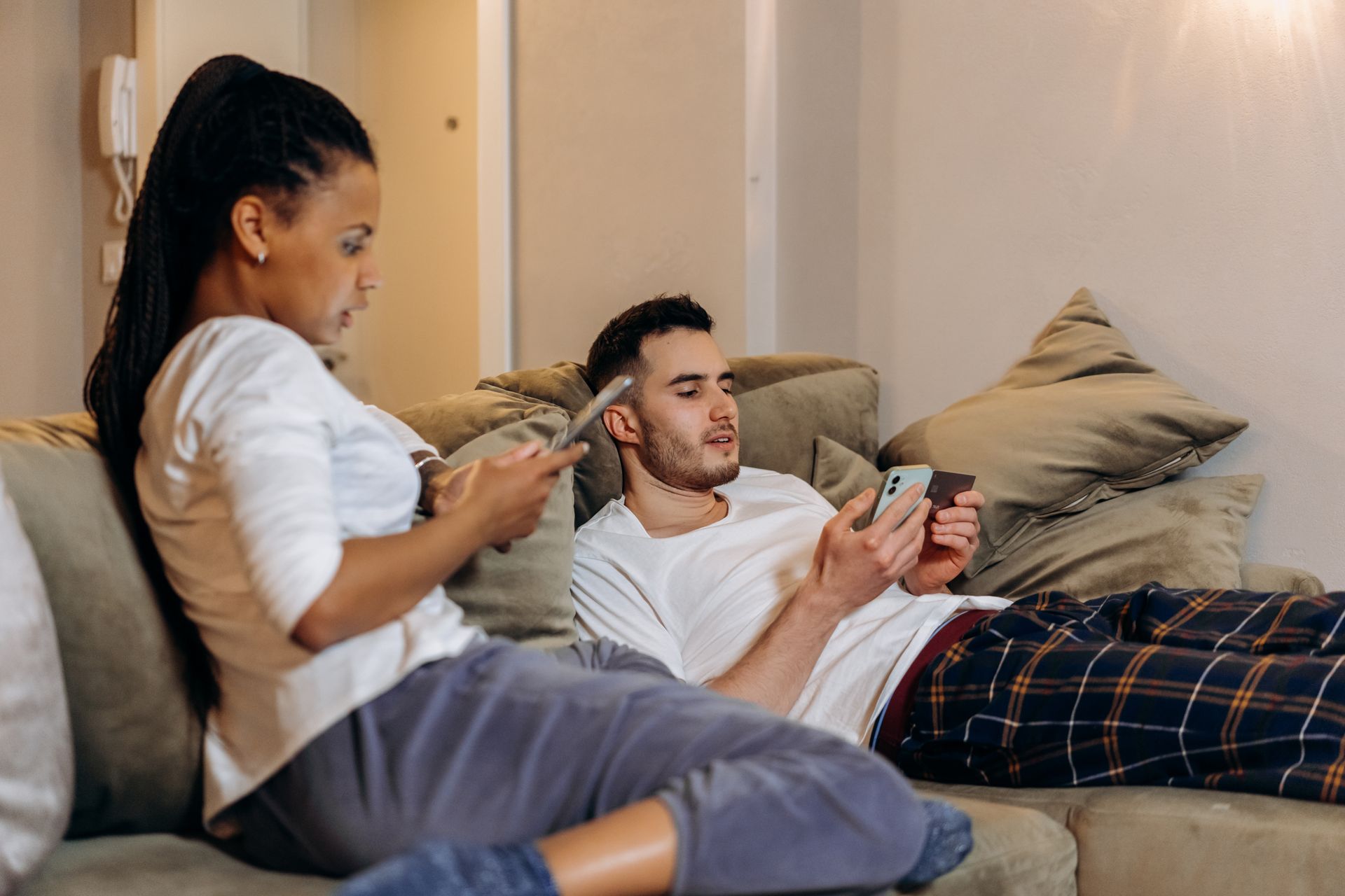 A couple using smartphones and a credit card for online shopping at home on the sofa.